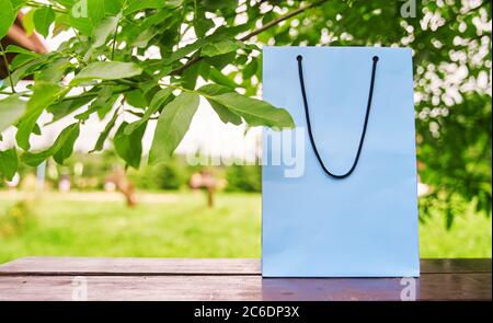 Blank paper bag stands on a wooden table against the background of green leaves. Shopping concept. Copy space. Stock Photo