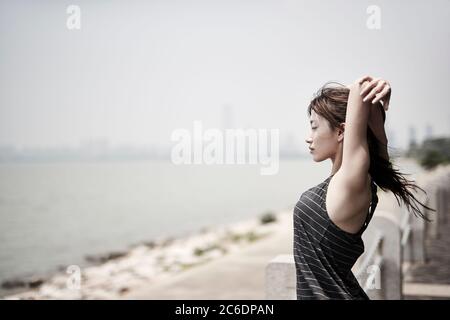 young asian adult woman stretching arms outdoors by the sea, side view