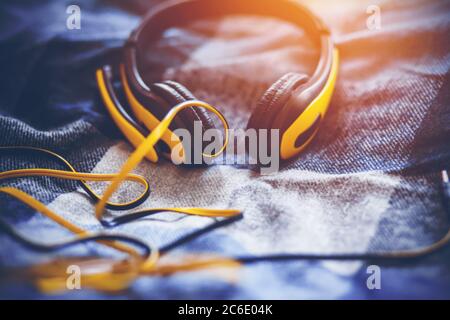 Fashionable yellow large headphones with a microphone and a long tangled wire lie on a soft plaid blanket on the bed and are illuminated by sunlight. Stock Photo