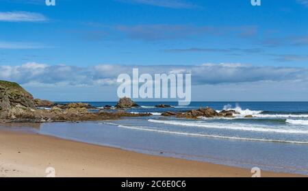 CULLEN BAY BEACH AND TOWN MORAY COAST SCOTLAND A BLUE SKY AND A SANDY BEACH WITH WAVES BREAKING ON THE ROCKS Stock Photo