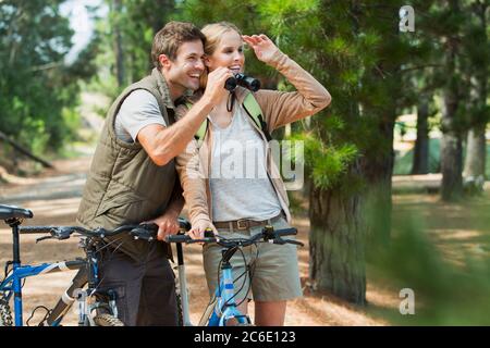Smiling couple with mountain bikes using binoculars in woods Stock Photo
