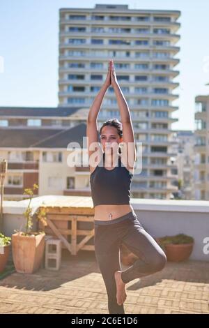 Young woman practicing yoga tree pose on sunny urban rooftop Stock Photo