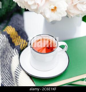 Cup of tea on books, flowers white wild rose in vase , warm plaid on white table outside in garden. Romantic leisure breakfast with nature background Stock Photo