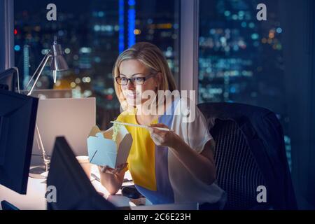 Businesswoman eating take out food at desk in office at night Stock Photo