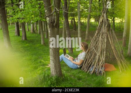 Woman with laptop relaxing at branch teepee in woods Stock Photo