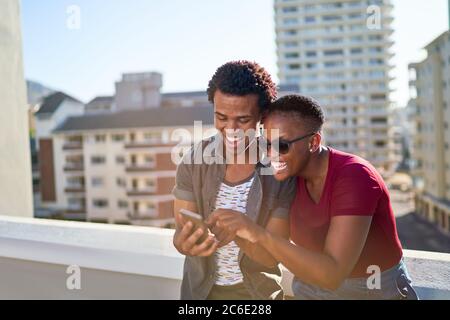Happy young couple using smart phone on sunny urban rooftop balcony Stock Photo