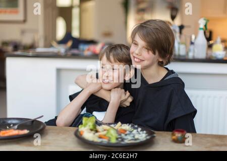 Happy brothers hugging and eating dinner at dining table Stock Photo