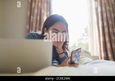 Teenage girl using smart phone at laptop on bed