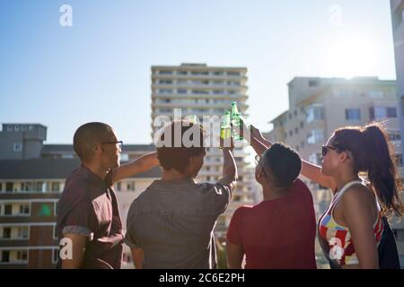 Young friends toasting beer bottles on sunny urban rooftop balcony Stock Photo