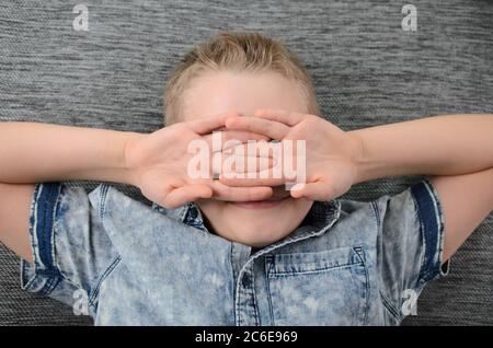 Cute funny boy is lying his backs on couch and making funny face, playful child boy having fun at home on sofa. Stock Photo