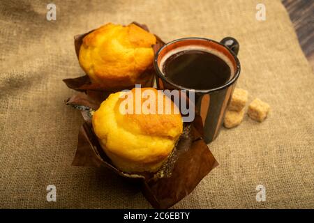 Two fresh cupcakes, a mug of coffee, and chunks of brown cane sugar on a rough-textured homespun cloth. Close up Stock Photo