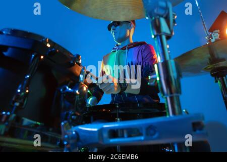Close up. Young inspired and expressive musician, drummer performing on gradient colored background in neon light. Concept of music, hobby, festival, art. Joyful artist, colorful, bright portrait. Stock Photo