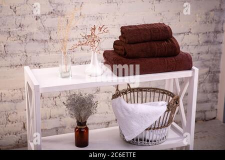 Rack with a stack of three brown color towels and baskets with clean white towels and toilet decor near a brick wall. Shelf with terry towels and Stock Photo