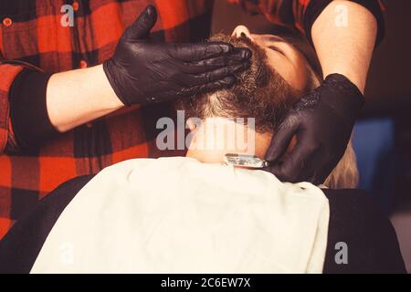 Hand of barber keeping straight razor and cutting trendy style on beard of client. Concept of shaving. Stock Photo