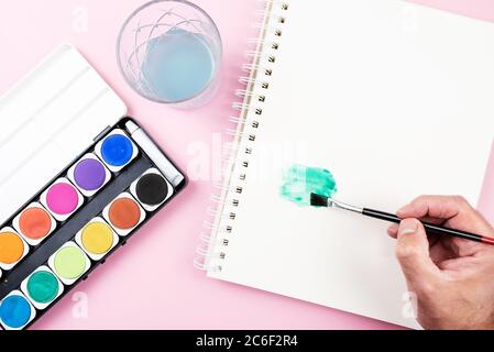 painting with watercolors, top view of paintbox, sketch pad and person using paintbrush Stock Photo