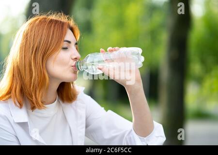 Happy thirsty redhead woman drinking fresh bottled water in summer outdoors. Stock Photo