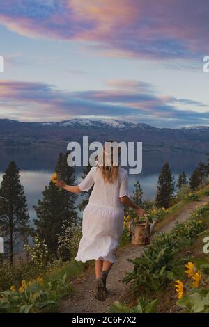 Woman  in white dress and blond hair running on path with  watering can Stock Photo