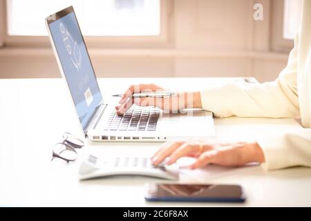 Close-up shot of businesswoman’s hand typing on laptop and using calculator while working at office desk. Home office. Stock Photo