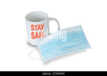 A light blue medical face mask in front of a white mug with red text saying STAY SAFE on white background. Prevention of Coronavirus Covid-19 concept. Stock Photo