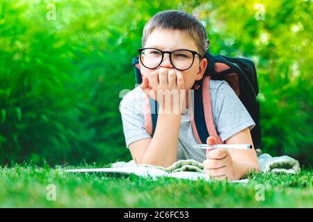 thoughtful kid boy in glasses thinking of solution. Funny schoolboy looking for idea answer to question. Young happy toddler laying on grass with back Stock Photo