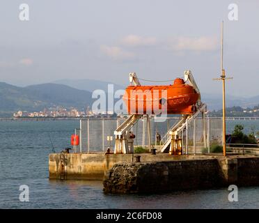 Bright orange decommissioned capsule life boat suspended in a white launch frame next to the bay in Santander Cantabria Spain Stock Photo