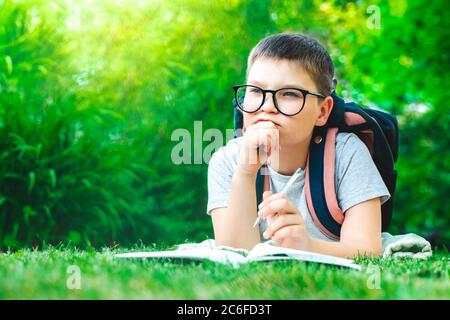 thoughtful schoolboy in glasses thinking of solution, idea. dreamy child boy answering question. Young creative toddler laying on grass with backpack Stock Photo