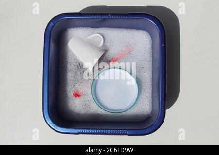 A dishwashing brush, a cup and a bowl in a blue plastic washing-up bowl with soapy water. Stock Photo