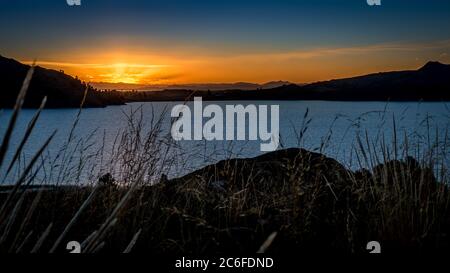 sunset with an orange and dark blue cloudy sky and sunbeams on lake titicaca, grasses in the foreground with an extensive landscape and fog in valley Stock Photo
