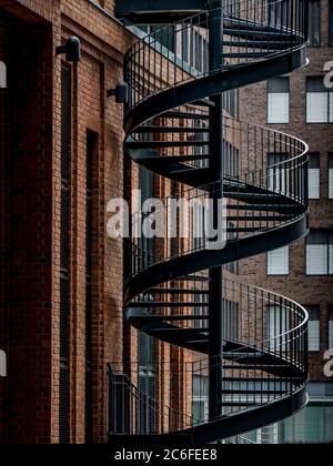 frontal view of steel spiral staircase in industrial design leading upwards next to a brick facade in a modern backyard outside of an office building Stock Photo