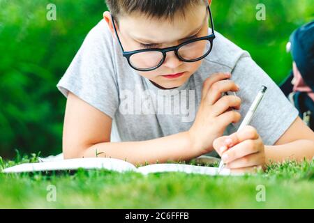 portrait of studying schoolboy in glasses laying on grass writing in exercise book making homework. male child makes notes does math outdoors in park. Stock Photo