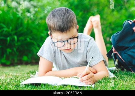 Concentrated schoolboy in glasses laying on grass writing in exercise book making homework. male child drawing geometric figures writing notes doing m Stock Photo