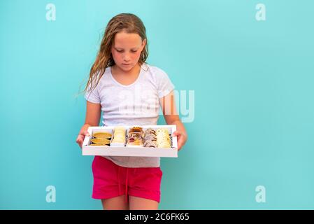 Cute teenage girl freckles woman holding box with dessert chocolate and vanilla mousse decorated with sweet cream, cookies and candy in a glass jar, fruit sushi, alfajores, macaroons and creme brulee. Stock Photo