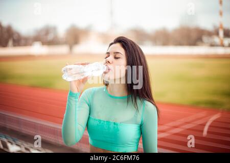 Beautiful girl drinks water from a bottle after sports training. Girl in an aquamarine topic with her hair against the backdrop of the stadium. Stock Photo