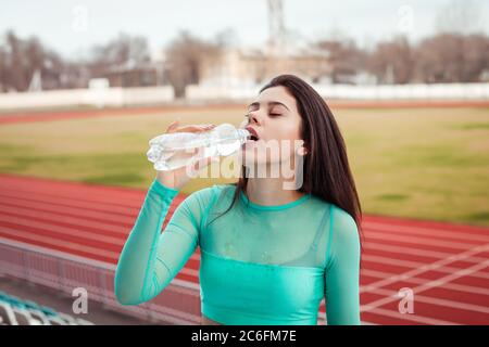 Beautiful girl drinks water from a bottle after sports training. Girl in an aquamarine topic with her hair against the backdrop of the stadium. Stock Photo