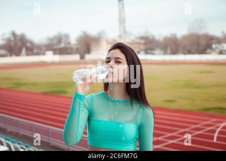 Beautiful girl drinks water from a bottle after sports training. Girl in an aquamarine topic with her hair against the backdrop of the stadium. Stock Photo