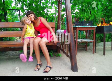 Mom and daughter cuddle cheerfully. Little girl with mom laugh fun in the garden. Family gatherings on the background of a bonfire in nature. Stock Photo