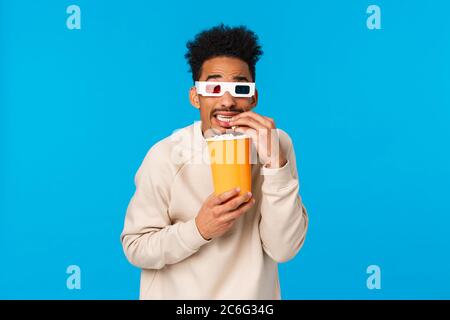 So scarry but interesting. Enertained handsome funny african american guy watching horror movie in cinema, eating popcorn and cringe looking screen Stock Photo