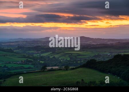 Askerswell, Dorset, UK.  9th July 2020.  UK Weather.  View from Eggardon Hill near Askerswell in Dorset looking towards the hills of Pilsdon Pen and Lewesdon Hill as a break in the dark moody clouds glow orange at sunset.  Picture Credit: Graham Hunt/Alamy Live News Stock Photo