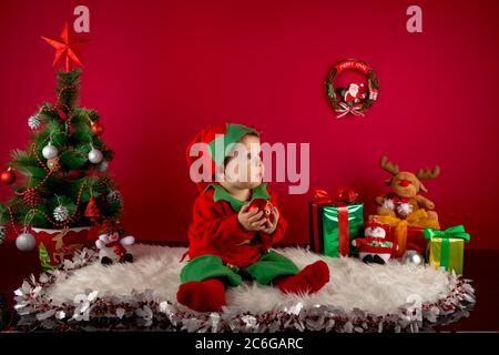 Baby dressed as an elf on her first Christmas, set in a setting with a Christmas tree, gifts, snow, reindeer and decorations Stock Photo