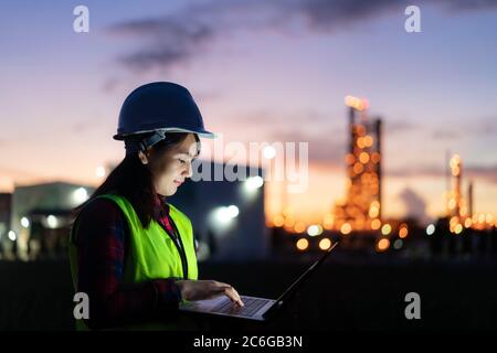 Asian woman petrochemical engineer working at night with laptop Inside oil and gas refinery plant industry factory at night for inspector safety quali Stock Photo