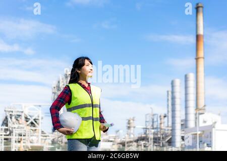 Asian woman engineer holding a white safety helmet and blueprint looking forward to future with oil refinery plant factory in background. Stock Photo