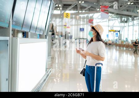 Asian woman tourist wearing face mask checking flight from arrival departure board in airport terminal for new normal travel concept. Stock Photo