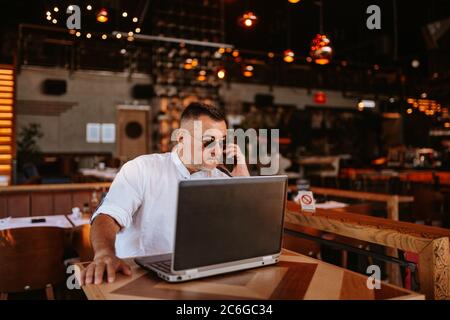 A middle-aged caucasian businessman with glasses in a white shirt is sitting in a cafe, talking on the phone and working on a laptop Stock Photo