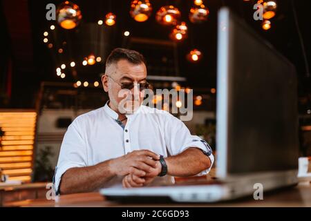 A middle-aged caucasian man with glasses in a white shirt sitting in a cafe looking at his watch while a laptop is on the table Stock Photo