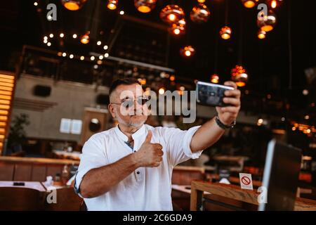 A middle-aged caucasian businessman with glasses in a white shirt sits in a cafe and takes a selfie while a laptop is on the table Stock Photo