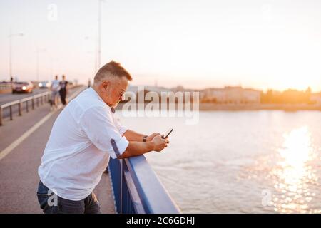 A middle-aged caucasian businessman with glasses, in a white shirt, is typing a message on the phone leaning against the bridge railing. Stock Photo