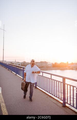 A middle-aged caucasian  businessman with glasses, in a white shirt and with a black briefcase  in his hand walks on the bridge and looks at his watch Stock Photo
