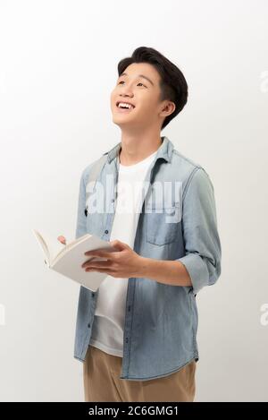 A young Asian man standing in a blue shirt, holding a book and reading, isolated for white background. Stock Photo