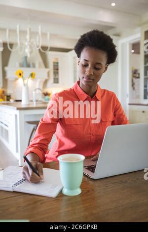 Woman taking notes while using laptop in the kitchen Stock Photo