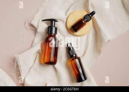 Amber glass cosmetic packaging set without labels. Flat lay pump bottle dispenser, dropper bottle and spray on beige fabric. Top view, overhead. Beaut Stock Photo